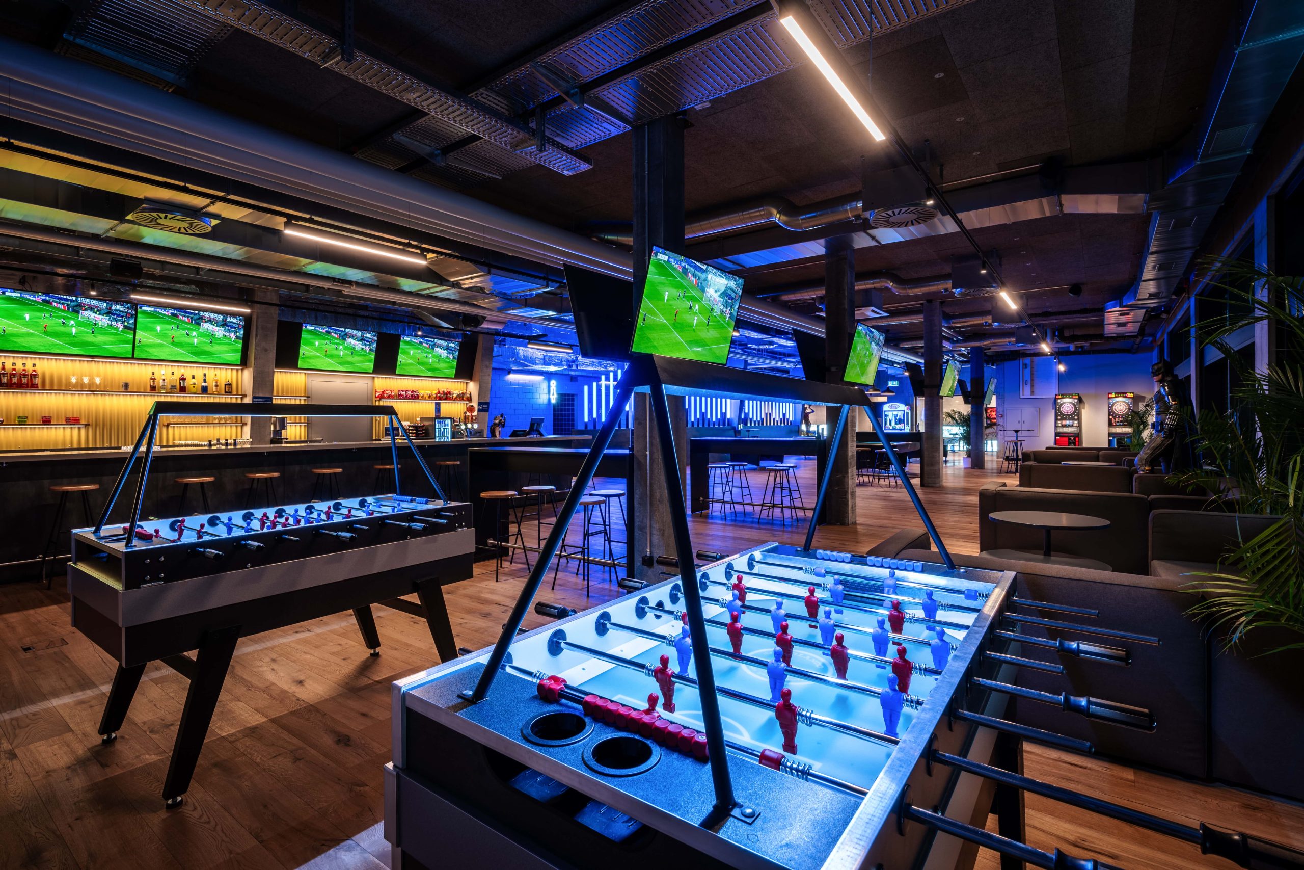 Vibrant game zone at blue Cinema with foosball and pool tables amidst sports broadcasts.